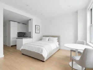 Minimalistisch eingerichtetes Apartment mit greigem Bettgestell, rundem Tulpen-Esstisch und hellem Eichenholzboden.