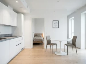 Minimalistisch eingerichtetes Apartment mit hellem Parkettboden, weißer Küche, rundem Tulpen-Esstisch und Blick ins schlichte Schlafzimmer.