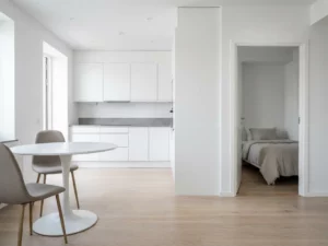 Minimalistisch eingerichtete Wohnung mit hellem Parkettboden, weißer Einbauküche, grauer Arbeitsplatte und rundem Tulpen-Esstisch mit greigen Polsterstühlen.