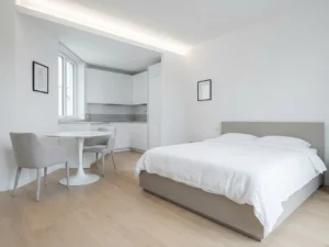 Minimalistisch eingerichtetes Apartment mit greigem Bettgestell, rundem Tulpen-Esstisch und weißer Einbauküche auf hellem Eichenparkett.