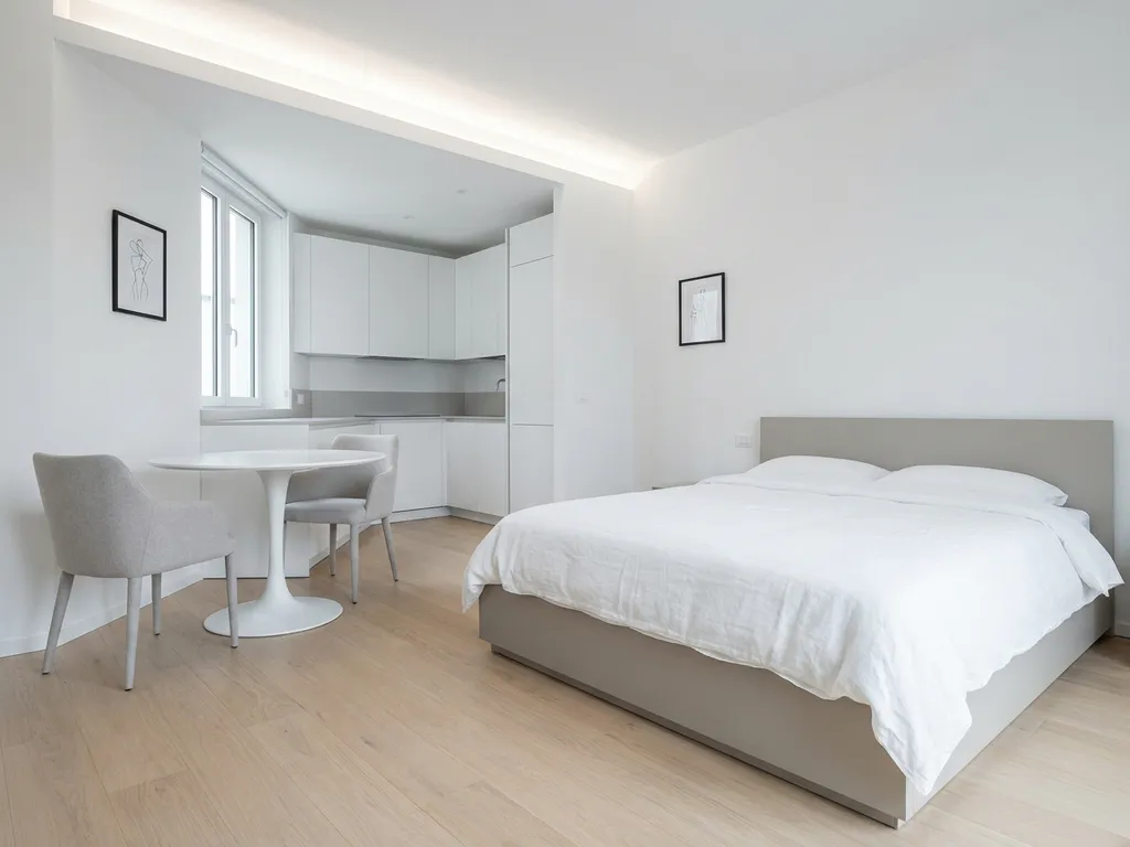 Minimalistisch eingerichtetes Apartment mit greigem Bettgestell, rundem Tulpen-Esstisch und weißer Einbauküche auf hellem Eichenparkett.