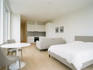 Minimalistisch eingerichtetes Apartment mit Greige-Bett, rundem Tulpen-Esstisch, hellem Holzboden und weißen Küchenschränken.