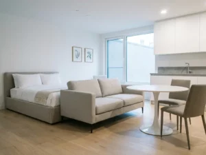 Minimalistisch eingerichtetes Apartment mit greigefarbenem Bett, Sofa, weißer Tulpen-Esstisch und hellem Eichenholzboden.