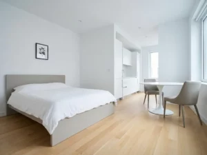 Minimalistisch eingerichtetes Apartment mit greigefarbenem Bettrahmen, rundem Tulip-Esstisch und hellen Holzböden.