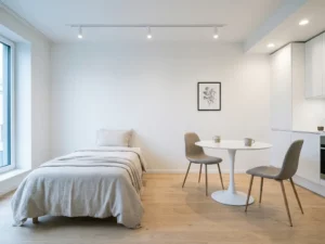 Minimalistisch eingerichtetes Apartment mit Einzelbett, rundem Tulip-Esstisch und weißen Küchenschränken auf hellem Eichenparkett.