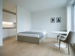 Minimalistisch eingerichtetes Apartment mit greigem Bettgestell, rundem Tulpen-Esstisch und weißer Einbauküche auf hellem Parkettboden.