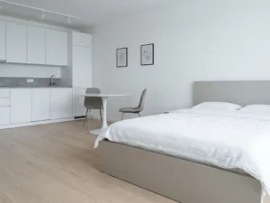 Minimalistisch eingerichtetes Apartment mit greigefarbenem Bett, rundem Tulpen-Esstisch und hellem Eichenholzboden.