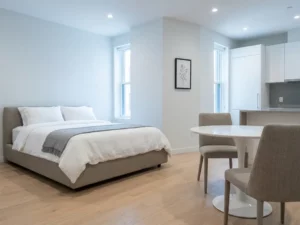 Minimalistisch eingerichtetes Apartment mit greigem Bettgestell, rundem Tulpen-Esstisch und hellen Eichenholzdielen.
