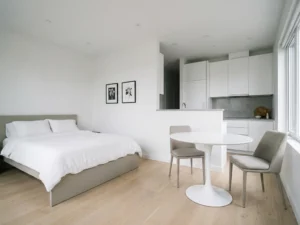 Minimalistisch eingerichtetes Apartment mit greigefarbenem Bett, rundem Tulpen-Esstisch und hellem Eichenholzboden.