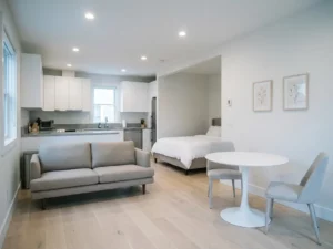 Minimalistisch eingerichtete Wohnung mit greigem Bett, Zweisitzer-Sofa, rundem Tulpen-Esstisch und hellen Holzdielen.