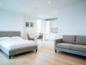 Minimalistisch eingerichtetes Apartment mit greigem Bettgestell, Zweisitzer-Sofa, rundem Tulpen-Esstisch und hellem Eichenparkett.
