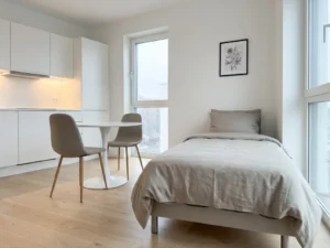Minimalistisch eingerichtetes Apartment mit greigem Einzelbett, rundem Tulpen-Esstisch und hellen Holzböden.