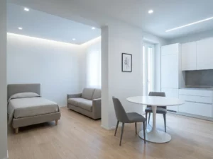 Minimalistisch eingerichtete Wohnung mit Tulip-Esstisch, Greige-Sofa, hellen Holzböden und weißen Küchenschränken.