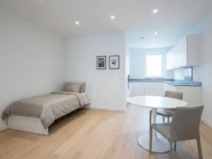 Minimalistisch eingerichtetes Apartment mit Einzelbett, rundem Tulip-Tisch und hellen Holzböden in Weiß- und Greigetönen.