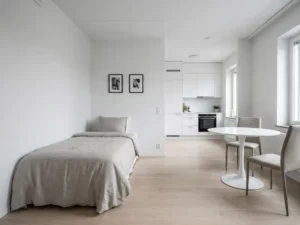 Minimalistisch eingerichtetes Apartment mit Einzelbett, rundem Tulip-Esstisch und weißer Einbauküche auf hellem Parkettboden.