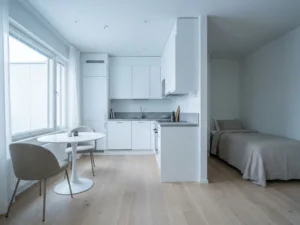 Minimalistisches Studio-Apartment mit hellem Parkettboden, weißer Küche, grauer Steinarbeitsplatte und rundem Tulpen-Esstisch.