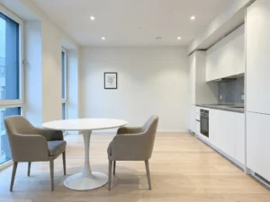Minimalistisches Apartment mit hellem Parkettboden, weißer Einbauküche, grauer Steinarbeitsplatte und rundem Tulpen-Esstisch mit zwei greigen Stühlen.