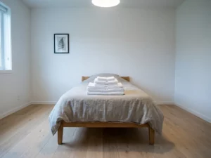 Gefaltete weiße Bettwäsche und Handtücher auf einer greige Bettdecke in einem minimalistischen Schlafzimmer mit hellem Holzboden.