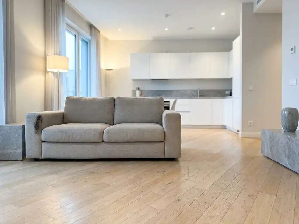 Zerkratzter hellblonder Parkettboden neben einem graubeige Zweisitzer-Sofa mit Scheuerstelle am Armlehnenbezug in minimalistischer Wohnung.